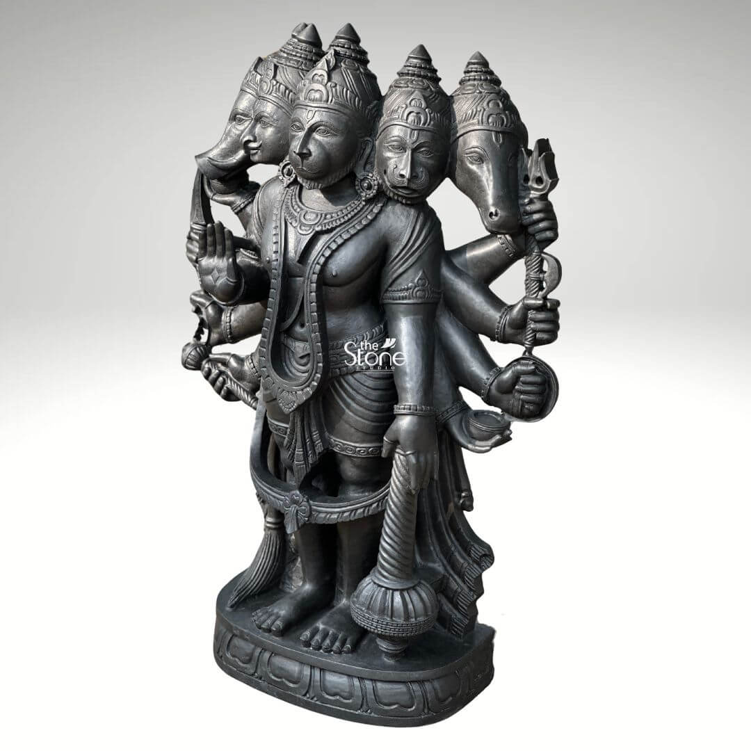 A three-quarter view studio photograph set against a plain light grey background displays a magnificent 3.5-foot Panchmukhi Hanuman Stone Murti. This intricately hand-carved Vigraham is made from a single block of deep dark blackstone renowned for its durability and fine grain. The statue depicts the five-faced form of Lord Hanuman known as Panchamukhi Hanuman standing firmly on a multi-tiered ornate circular base. The central face is that of Hanuman himself looking forward with a serene protective expression adorned with traditional jewelry and a crown. Radiating outwards from the central head are four other divine faces: to the east the boar face to the south the lion face to the west the eagle face and to the north the horse face. The statue is multi-armed totaling ten arms that hold various divine attributes each rendered with exquisite detail. The main central right hand is raised in the Abhaya mudra while the main central left hand holds a mace that rests on the base. The other eight hands hold a conch discus sword shield noose hook a mountain and a vessel. The deity is adorned with elaborate traditional South Indian carvings of necklaces armlets wristlets a sacred thread and a detailed dhoti all chiseled with precision into the dark stone. The fine texture of the blackstone enhances the depth and shadow of the carvings highlighting the complex iconography of this formidable yet benevolent form of Hanuman. This Panchmukhi Hanuman Stone Murti is an exceptional example of traditional Indian temple sculpture, suitable for sanctum placement or as a grand focal point in a home shrine or spiritual space. The 3.5-foot height makes it a commanding presence embodying the combined power of the five deities represented by the faces. The blackstone material gives the statue a timeless ancient feel ensuring it will be a cherished piece for generations. The statue's detailed iconography from the expressions on the five faces to the specific items held in the ten hands, makes it a significant subject for study and devotion. The expert craftsmanship transforms the raw stone into a vibrant sacred form.
