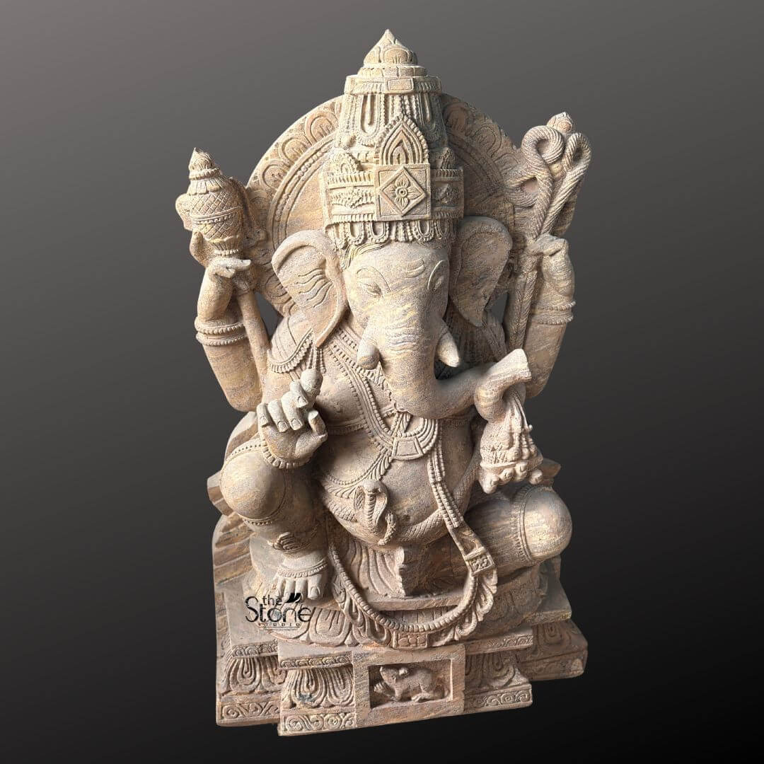 A detailed eye-level photograph of a magnificent Pillaiyar Stone Statue hand-carved from a single block of warm brown sandstone. The statue stands approximately four feet tall and is depicted in a traditional seated posture known as lalitasana, with one leg gracefully folded and the other resting on a decorated base. The four-armed deity is seated upon a carved multi-tiered lotus pedestal which is further embellished with intricate floral and geometric patterns and features a small detailed carving of Ganesha's vehicle the mouse centrally at the base. The sandstone exhibits a natural slightly porous texture and warm color variations typical of the material showcasing the artisan's skill in achieving fine detail. Pillaiyar or Lord Ganesha is richly adorned with abundant deeply carved traditional jewelry and ornaments all rendered in the same sandstone. He wears a detailed jeweled mukuta featuring a prominent central gem and peacock motifs multiple necklaces of varying lengths elaborate armlets wristlets and anklets. His sacred thread is intricately carved across his torso. The deity holds symbolic attributes in his four hands his upper right hand holds an axe and his upper left hand holds a noose while his lower right hand is raised in a gesture of blessing and his lower left hand holds a sweet towards his trunk. The trunk itself curves elegantly to the left holding the modak. This commanding 4 feet statue embodies the spiritual essence and iconographic details of the remover of obstacles making it a suitable and sacred focal point for a home shrine or temple sanctum. The face of Pillaiyar is serene and benevolent with downcast eyes conveying deep wisdom and compassion. The careful carving of every element from the tassels of his garments to the petals of the lotus throne reflects centuries-old sculpting traditions. The overall composition is balanced and reverent highlighting the divine form of Lord Ganesha. The photograph captures the statue against a plain dark grey studio background emphasizing the detailed craftsmanship and the natural beauty of the sandstone.