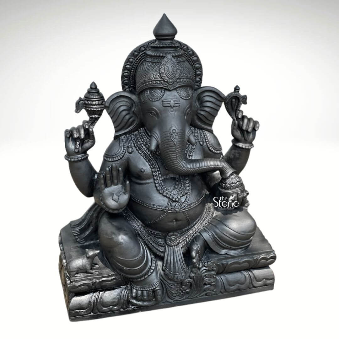 A three-quarter view of a magnificent 2.5ft Pillaiyar Stone Statue meticulously carved from a single piece of deep black stone. Seated in the traditional lalitasana posture Lord Ganesha is depicted with four arms adorned in intricate deeply carved jewelry that highlights the dark stone's matte finish. His upper right hand holds a parashu and his upper left holds a pasha both symbols of power. The lower right hand is raised in the abhaya mudra while the lower left hand cradles a modaka his trunk curving gracefully to taste it. The deity's form is substantial and rounded seated upon a tiered pedestal carved to resemble stacked lotus petals which provides a stable base for the statue. A small intricately carved mooshika his vehicle is nestled at the base of the pedestal near his left foot looking up in devotion. The fine detailing on the crown the sacred thread the mala beads and the folds of his dhoti demonstrates exceptional craftsmanship capturing the divine elegance and rich symbolism associated with Pillaiyar. This Blackstone Ganesha statue is a striking example of traditional Indian stonework embodying the spiritual significance central to an article about Pillaiyar Stone Statues. The dark color of the basalt stone imparts a sense of solemnity and ancient heritage making it a commanding presence. The texture and craftsmanship visible in this image are characteristic of premium stone art showcasing the divine form of the remover of obstacles as a masterful work of sculpture.