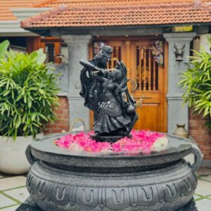 A magnificent 3.5-foot-tall Radha Krishna statue meticulously carved from dark polished sandstone stands as the majestic centerpiece at the entrance of a traditional Indian home in Thiruvananthapuram. The divine couple is depicted in a graceful loving pose with Krishna playing the flute and Radha gazing up at him adoringly their forms showcasing intricate detailing in the sculpting of their traditional attire jewelry and crowns. The statue is positioned atop a large ornate dark granite stone base that features carved U-shaped patterns adding to its grandeur and grounded presence. The statue is nestled within a circular shallow water feature brimming with a vibrant bed of fresh pink and white lotus flowers and floating petals which creates a lush fragrant setting and enhances the spiritual atmosphere of the entrance. The water element symbolizes purity and devotion beautifully complementing the sandstone sculpture. The placement of the idol at the home entrance visible against the backdrop of a traditional Kerala home with a tiled roof wooden door and large potted plants like bamboo and palms provides a perfect example of integrating sacred art into home decor. This image illustrates a harmonious and culturally rich way to decorate a home entrance offering inspiration for those seeking to install a Radha Krishna stone statue for a similar purpose in Thiruvananthapuram. The 3.5-foot size is ideal for commanding attention without overwhelming the space serving as a focal point that welcomes guests with a sense of peace and divinity. The durable sandstone material is well-suited for the tropical climate of Kerala ensuring the statue remains a beautiful and revered part of the home for years to come embodying the essence of devotion and artistic elegance.