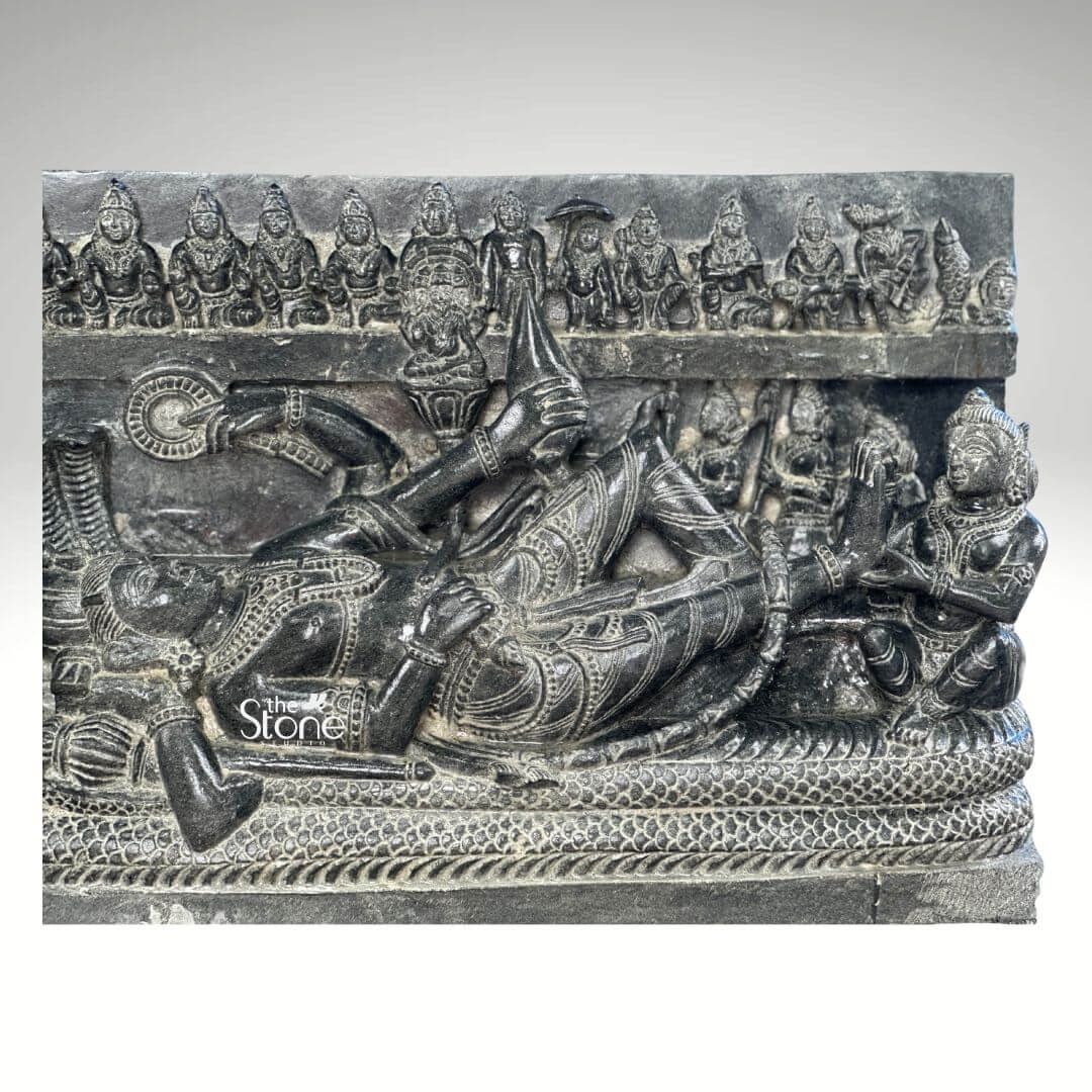 A detailed hand-carved black stone statue approximately 30 inches tall depicting the Hindu deity Lord Vishnu in the Anantashayana posture reclining upon the cosmic serpent Sheshnag. The central figure of Vishnu lies peacefully on the coiled body of the multi-hooded serpent whose seven hooded heads fan out to form a protective canopy above the deity's crowned head. Vishnu is adorned with traditional jewelry a tall crown and elaborate garments with his right hand gently supporting his head while his left arm rests along his body holding a conch shell. The sculpture is carved in high relief showcasing the rich dark matte texture of the blackstone. Below the main reclining figure the coils of Sheshnag are intricately detailed forming a textured bed. Several attendant figures are carved into the stele including a seated Goddess Lakshmi massaging his foot at the base. Above the serpent canopy a frieze of miniature figures representing the ten avataras of Vishnu is carved horizontally across the top of the panel adding a comprehensive narrative to the divine iconography. The sculpture exhibits a refined finish with the natural variations in the stone's color and texture conveying a sense of ancient artistry and reverence consistent with a high-quality stone statue. The plain neutral grey background isolates the form and draws the viewer's eye directly to the complex narrative and divine iconography depicted in the stone. This detailed carving serves as an exemplary illustration for an article discussing reclining Lord Vishnu black stone statues.