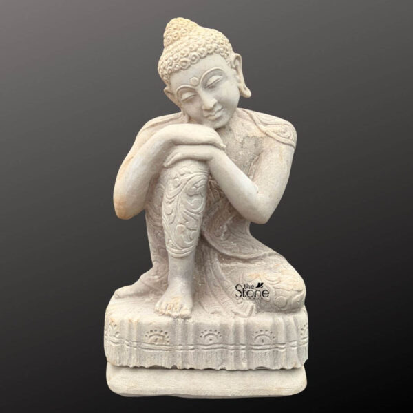 A three-quarter view studio photograph of a 16-inch white sandstone statue depicting the Resting Buddha designed for garden use. The statue is hand-carved from a light-colored fine-grained sandstone exhibiting a natural matte texture and a serene contemplative expression. The Buddha is seated in a relaxed asymmetrical posture with his left knee raised and his right leg crossed in a loose half-lotus position. His head with characteristic snail-shell curls and a prominent ushnisha is tilted gently forward resting peacefully upon his clasped hands which are placed atop his raised left knee. The figure wears flowing elegantly draped robes that reveal his shoulders and are subtly textured with decorative carved floral patterns adorning the edges of the fabric and the front of his folded legs. The skin is smooth and serene with downcast eyes conveying deep tranquility. The statue is positioned on a rectangular sandstone base which is carved with a repeating stylized floral and leaf motif across its front face. The base anchors the figure and provides stability making it suitable for placement on garden pedestals pathways or amidst foliage. The overall color of the statue is a warm creamy white with subtle natural variations and speckling inherent to the sandstone material. This light neutral hue allows the statue to stand out against the greenery of a garden creating a focal point of peace and reflection. The natural lighting highlights the delicate carving of the robes the texture of the hair curls and the smooth contours of the Buddha's face emphasizing the craftsmanship and the meditative quality of the piece. The statue's pose and material make it a fitting addition to outdoor garden spaces seeking a touch of spiritual calm.