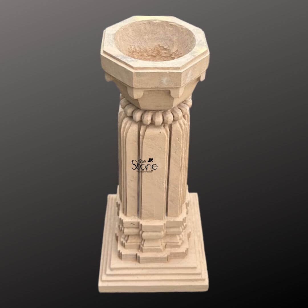 A three-quarter view photograph captures a magnificent Sandstone Tulsi Planter standing approximately 3 feet tall. This traditional Indian planter is hand-carved from a single block of warm brown sandstone exhibiting a natural textured finish. The entire structure rests upon a substantial stepped square base providing excellent stability and a sense of grounded grandeur perfect for a garden or courtyard setting. The main body of the planter features a beautifully proportioned square column adorned with classic vertical fluting on all four faces creating elegant lines that enhance its architectural presence. The sandstone surface displays subtle variations in color and grain highlighting the authentic artisanal quality of the hand-carving process. The precision of the work reflects a deep respect for traditional Indian stone carving techniques. Crowning the column is a beautifully shaped flared planter bowl designed to hold the sacred Tulsi plant and soil. This upper section transitions smoothly from the square column to a circular form featuring a decorative band of carved bead-and-reel molding just below the rim. The overall form is a harmonious blend of functionality and aesthetic grace making this sandstone Tulsi planter a revered and beautiful focal point for any home or garden.