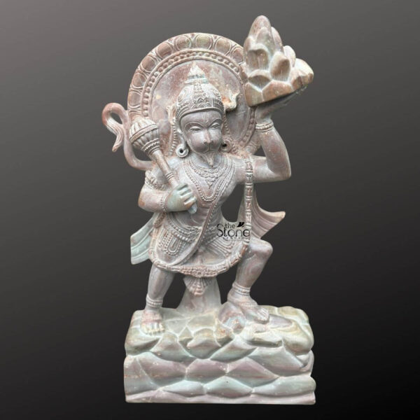 A detailed eye-level photograph of a handcrafted Sanjeevani Hanuman Stone Statue meticulously carved from a single block of pink soapstone. The statue stands approximately 2 feet tall capturing the god in a dynamic heroic pose balanced mid-air on a rocky mountain formation which represents Mount Dronagiri. Hanuman is depicted with a powerful yet compassionate expression looking intently forward. The natural color variations of the pink soapstone ranging from soft blushes to deeper hues are evident throughout the sculpture giving it a warm and unique texture. The statue is richly adorned with detailed carvings of traditional jewelry and ornaments all rendered in the soft stone. He wears an ornate crown multiple necklaces armlets wristlets and anklets with a sacred thread intricately carved across his torso. A flowing decorative cloth is draped around his waist and his tail is curled elegantly behind him forming part of the statue's support. The four-armed deity holds symbolic attributes: his upper right hand wields a mace while his upper left hand reverently holds the uplifted mountain peak the Sanjeevani Parvata complete with tiny carved medicinal herbs. His lower right hand is raised in a gesture of blessing and his lower left hand rests near his hip. This Sanjeevani Hanuman Stone Statue serves as a powerful representation of the Ramayana's pivotal moment when Hanuman fetches the mountain of life-saving herbs. The sculpture's form reflects centuries-old Indian temple art capturing the spiritual essence and heroic attributes of Lord Hanuman. The careful attention to anatomical detail from the musculature of the body to the texture of the fur showcases the master craftsmanship involved in working with pink soapstone. The overall composition conveys a profound sense of devotion and artistry making this statue a significant and beautiful addition to any home shrine or temple setting.