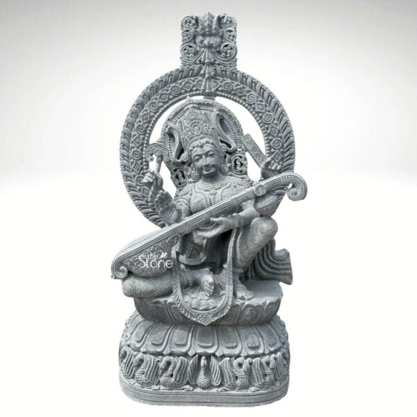 A full three-quarter view photograph of a magnificent Saraswati Devi Stone Vigraham hand-carved from a single block of dark textured black stone. The goddess of knowledge and arts is depicted in a graceful traditional seated posture known as lalitasana with one leg folded and the other resting on a lotus pedestal. She is seated upon an intricately carved multi-tiered padmasana base which is adorned with detailed petal motifs and a row of small carved figures along its front edge all rendered in the same deep black stone. The statue's surface exhibits a natural matte finish that highlights the rich dark color and the fine detail of the craftsmanship. Saraswati Devi is depicted with four arms holding symbolic attributes. Her primary hands are gracefully engaged in playing an elaborately carved veena a stringed musical instrument which rests across her lap. Her upper right hand holds a mala for meditation while her upper left hand holds a pustaka representing knowledge and wisdom. She is adorned with abundant finely detailed traditional South Indian jewelry including multiple necklaces elaborate armlets wristlets a jeweled mukuta and cascading garlands all intricately carved in high relief. A serene composed expression graces her face with downcast eyes conveying deep concentration and divinity. An ornate circular prabhavali frames the entire statue rising from behind the lotus throne. This decorative arch is embellished with intricate scrollwork floral patterns and a central kirtimukha motif at its apex. The entire vigraham from the base of the lotus throne to the top of the prabhavali stands 4.5 feet tall making it a substantial and commanding presence. The statue's form reflects the rich tradition of Indian temple art capturing the spiritual essence and iconographic details of Saraswati Devi. The careful carving of every element from the strings of the veena to the petals of the lotus showcases the master craftsmanship and the natural beauty of the black stone. This Saraswati Devi Stone Vigraham is a fine example of traditional Indian sculpture suitable for worship or as a significant decorative art piece.