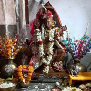 A detailed devotional photograph captures a seated Saraswati Stone Statue, approximately 3 feet tall and hand-carved from a warm brown sandstone. The statue rich in the natural texture of the stone is the central focus of a home puja altar. The goddess is depicted in a classic meditative pose seated on a lotus pedestal playing the veena held in her four arms. She is adorned with intricate carvings of traditional jewelry garlands of fresh white flowers, and a vibrant red and gold-bordered cloth draped over her shoulders. The statue is nestled within a sacred domestic space, surrounded by offerings for the festival of Saraswati Puja. To the left and right large brass pots or ghatas hold elaborate arrangements of colorful artificial flowers creating a festive backdrop. In the foreground on the floor are traditional ritual items: a bowl of puffed rice various fruits a small brass pot with a coconut and marigold garland and tiny metal cups. A simple alpana design is drawn in white powder on the stone floor beneath the altar grounding the statue in its cultural context. The scene is illuminated by soft warm indoor light highlighting the gentle serene expression on the goddess's face and the detailed craftsmanship of the sandstone carving. The natural imperfections and warm tones of the stone convey a sense of antiquity and deep reverence. The setting with its personal lived-in feel illustrates how such stone statues are venerated in households across Howrah serving as a tangible link to tradition and spirituality. The image is a perfect representation for an article discussing Saraswati stone statues and their place in the cultural life of the region.
