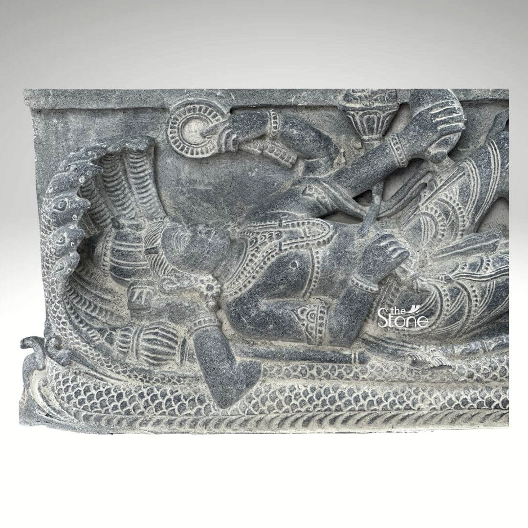 A detailed close-up photograph of a 30-inch blackstone sculpture depicting Lord Vishnu in the Sheshashayi pose reclining upon the cosmic serpent Sheshnag. The central figure of Vishnu lies peacefully his head supported by his right hand and resting upon the multiple hooded heads of Sheshnag, which form a protective canopy. He is adorned with intricate carvings of traditional jewelry including elaborate necklaces, armlets waistbands, and a tall cylindrical crown all rendered with high precision in the rich dark stone. His left arm rests along his body holding a conch shell. The sculpture is carved in high relief showcasing the deep matte texture of the blackstone. Below the reclining Vishnu the coils of Sheshnag are intricately detailed forming a textured bed that spans the length of the figure. Several attendant figures are carved into the stele including a seated Goddess Lakshmi massaging his foot at the base. The entire piece exhibits a refined finish with the natural variations in the stone's color and texture conveying a sense of ancient artistry and reverence consistent with a high-quality stone statue. The sculpture is presented against a plain neutral grey background which isolates the form and draws the viewer's eye directly to the complex narrative and divine iconography depicted in the stone. The detailed carving serves as an exemplary illustration for an article discussing Sheshashayi Lord Vishnu stone sculptures providing a clear view of the iconography and craftsmanship. The composition emphasizes the central reclining figure and the protective serpent hoods making it highly informative and accessible.