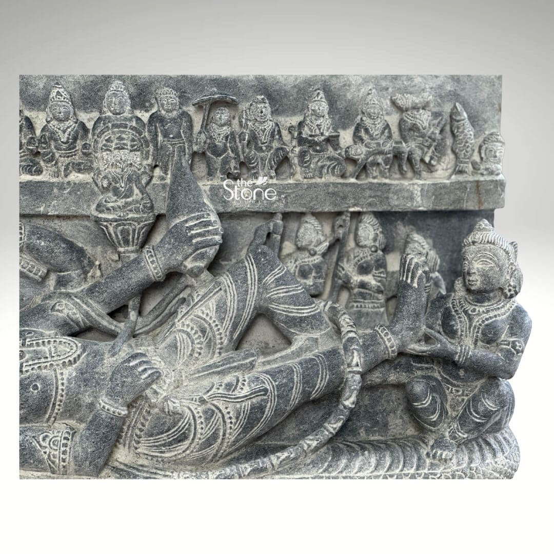 A detailed close-up photograph of a 30-inch blackstone sculpture depicting Lord Vishnu in the Sheshashayi pose, reclining upon the cosmic serpent Sheshnag. The central figure of Vishnu lies peacefully his head supported by his right hand and resting upon the multiple hooded heads of Sheshnag which form a protective canopy. He is adorned with intricate carvings of traditional jewelry including elaborate necklaces armlets waistbands and a tall cylindrical crown all rendered with high precision in the rich dark stone. His left arm rests along his body holding a conch shell. The sculpture is carved in high relief showcasing the deep matte texture of the blackstone. Below the reclining Vishnu the coils of Sheshnag are intricately detailed forming a textured bed. Several attendant figures are carved into the stele including a seated Goddess Lakshmi massaging his foot at the base and a frieze of miniature figures representing the ten avataras of Vishnu carved horizontally across the top of the panel adding a comprehensive narrative to the divine iconography. The sculpture exhibits a refined finish with the natural variations in the stone's color and texture conveying a sense of ancient artistry and reverence consistent with a high-quality stone statue. The plain neutral grey background isolates the form and draws the viewer's eye directly to the complex narrative and divine iconography depicted in the stone. The detailed carving serves as an exemplary illustration for an article discussing Sheshashayi Vishnu stone sculptures providing a clear view of the iconography and craftsmanship. The composition focuses on the lower body and feet of the reclining deity and the attendant goddess.