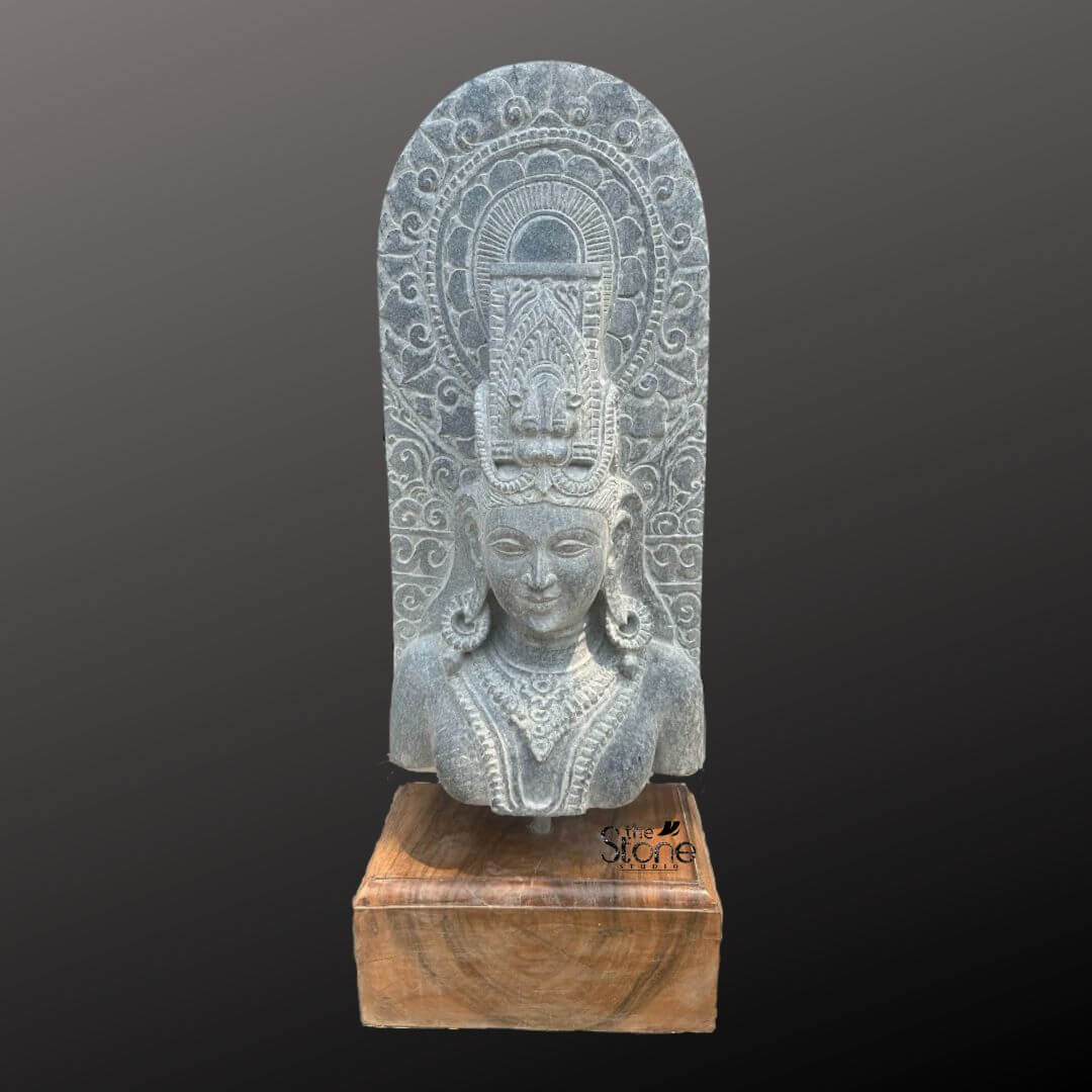 A magnificent thirty-inch hand-carved Lord Shiva bust in black stone stands as the revered centerpiece against a dark neutral gradient background. The sculpture meticulously chiseled from a single block of black stone exhibits a rich aged texture with natural variations and a weathered patina that underscores its antique character. Lord Shiva is portrayed with a serene contemplative facial expression characterized by eyes gently closed in meditation arched eyebrows and a subtle composed smile that conveys a profound sense of peace and divine wisdom. The bust features elaborate ornamentation including large circular earrings and multiple layered necklaces with intricate pendants that rest upon his chest all hand-carved with precision. His hair is intricately styled in a traditional jata forming a high crown adorned with delicate carvings of a crescent moon a serpent and a central floral motif with further details of coiled locks falling onto his shoulders. Behind the deity's head a richly carved ornate prabhavali rises featuring detailed, symmetrical floral and scrollwork patterns that frame his divine form. The craftsmanship showcases the traditional South Indian temple art style, with deep undercuts and high relief that create a profound sense of depth and reverence. The entire sculpture rests securely on a dark polished wooden plinth. This exquisite thirty-inch hand-carved Lord Shiva black stone bust is a perfect testament to the timeless devotion and artistic skill of traditional Indian artisans. The large scale of the bust combined with the deep black stone and the profound meditative expression makes it an awe-inspiring focal point for any spiritual space or home mandir. The detailed iconography from the elaborate headdress to the intricate jewelry and the ornate prabhavali ensures this antique-style murti meets the highest aesthetic and spiritual requirements for those seeking a significant and beautiful representation of Lord Shiva. The natural color of the stone and the profound depth of the carving convey a sense of sanctity and spiritual presence making it a lasting addition to any sacred environment.
