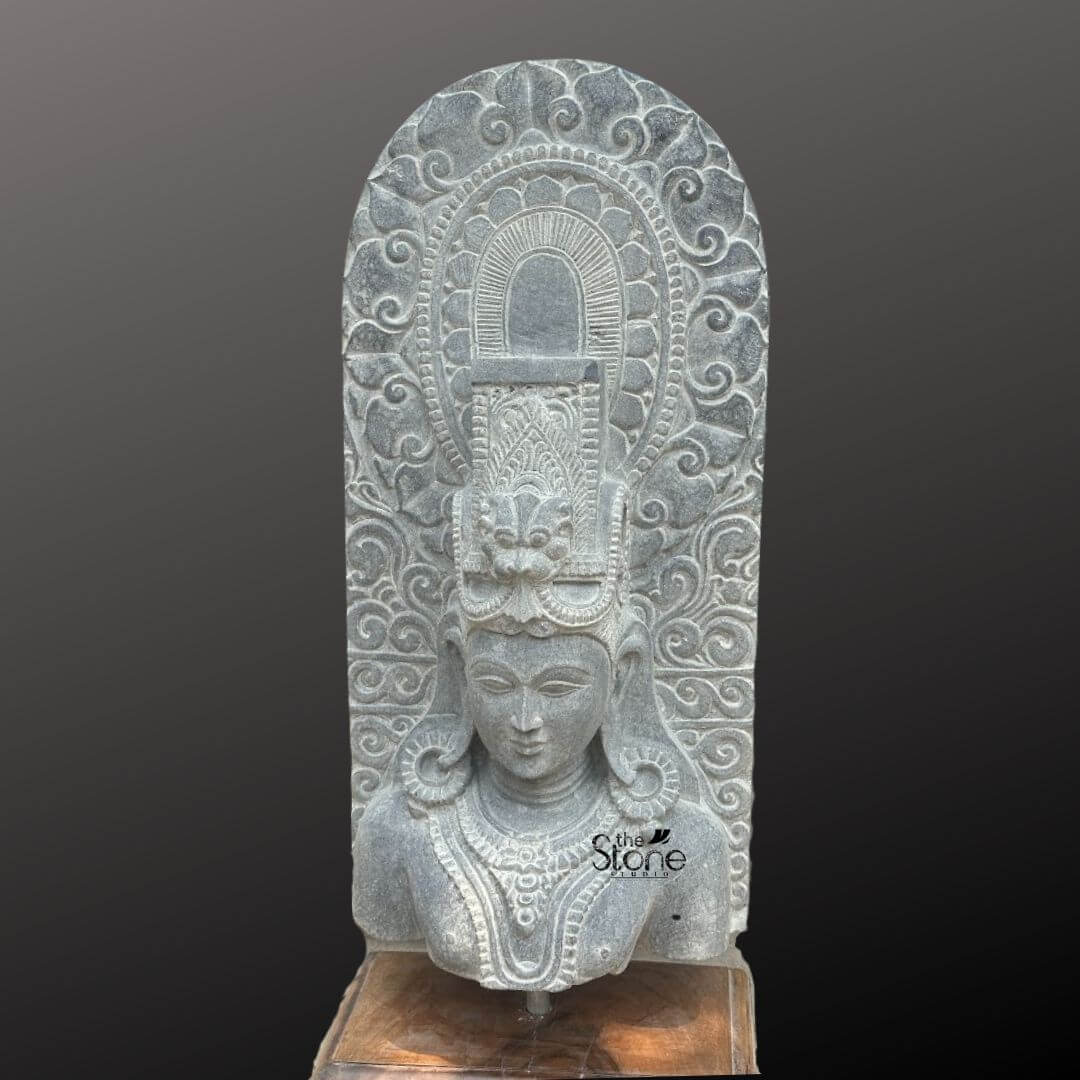 A magnificent thirty-inch hand-carved Lord Shiva bust in black stone stands as the revered centerpiece against a dark neutral gradient background showcasing the intricate sculptural details of the deity and the surrounding prabhavali. The sculpture meticulously chiseled from a single block of black stone exhibits a rich aged texture with natural variations and a weathered patina that underscores its antique character. Lord Shiva is portrayed with a serene contemplative facial expression characterized by eyes gently closed in meditation arched eyebrows and a subtle composed smile that conveys a profound sense of peace and divine wisdom. The bust features elaborate ornamentation including large circular earrings and multiple layered necklaces with intricate pendants that rest upon his chest all hand-carved with precision. His hair is intricately styled in a traditional jata forming a high crown adorned with delicate carvings of a crescent moon a serpent and a central floral motif with further details of coiled locks falling onto his shoulders. Behind the deity's head a richly carved, ornate prabhavali rises featuring detailed symmetrical floral and scrollwork patterns that frame his divine form. The craftsmanship showcases the traditional South Indian temple art style with deep undercuts and high relief that create a profound sense of depth and reverence. The entire sculpture rests securely on a dark polished wooden plinth. This exquisite thirty-inch hand-carved Lord Shiva black stone sculpture with a prominent prabhavali is a perfect testament to the timeless devotion and artistic skill of traditional Indian artisans. The large scale of the bust combined with the deep black stone the profound meditative expression and the ornate halo makes it an awe-inspiring focal point for any spiritual space or home mandir. The detailed iconography from the elaborate headdress to the intricate jewelry and the ornate prabhavali ensures this antique-style murti meets the highest aesthetic and spiritual requirements for those seeking a significant and beautiful representation of Lord Shiva. The natural color of the stone and the profound depth of the carving convey a sense of sanctity and spiritual presence, making it a lasting addition to any sacred environment.