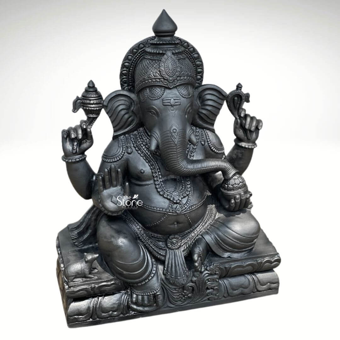 A detailed three-quarter view photograph of a magnificent Sitting Ganesha Black Stone Statue showcasing its divine form and intricate craftsmanship against a clean neutral background. The statue which stands approximately 2.5 feet tall is meticulously hand-carved from a single piece of deep dark black stone giving it a solemn and commanding presence. Lord Ganesha is depicted in a traditional relaxed seated posture known as lalitasana with his right leg folded and his left leg bent. The statue's surface is finished with a smooth dark polish that accentuates the natural texture of the stone and the precision of the detailed carving. Ganesha is adorned with elaborate deeply cut ornaments that cover his form including multiple necklaces armband wristlets, and anklets all rendered in the black stone. He wears a richly carved crown and a sacred thread across his chest. He has four arms; his upper right hand holds a parashu and his upper left holds a pasha. His lower right hand is raised in the abhaya mudra while his lower left hand cradles a mod which his gracefully curved trunk is poised to taste. The elephant head features fine details including traditional tilak markings on the forehead and large expressive ears. The statue is seated upon a substantial tiered pedestal also carved from the black stone which is designed to resemble stacked lotus petals. The base provides a stable and elegant foundation. At the lower left of the pedestal near Ganesha's foot a small intricately carved mooshika his vahana is depicted in a reverent pose looking up towards the deity. The overall composition highlights the divine elegance symmetry and rich iconography of this masterwork making it a quintessential example of a Sitting Ganesha Black Stone Statue suitable for spiritual spaces and art collections.