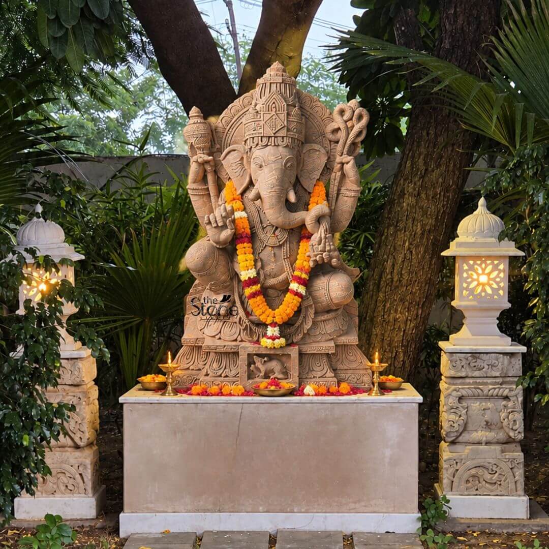 A serene eye-level photograph capturing a magnificent Sri Ganapathi Stone Idol meticulously hand-carved from a single block of warm brown sandstone. The statue stands approximately four feet tall and is depicted in a traditional seated posture known as lalitasana with one leg gracefully folded and the other slightly pendant. The four-armed deity is seated upon an intricately carved multi-tiered lotus pedestal which is further embellished with detailed floral and geometric patterns and features a small finely carved figure of Ganesha's vehicle the mouse centrally at its base. The sandstone exhibits a natural warm texture typical of the material showcasing the artisan's skill in achieving fine detail in the deity's form and ornaments. Sri Ganapathi is richly adorned with abundant deeply carved traditional jewelry and ornaments all rendered in the same sandstone. He wears a detailed jeweled mukuta featuring a prominent central gem and peacock motifs multiple necklaces of varying length elaborate armlets wristlets, and anklets. His sacred thread is intricately carved across his torso. The deity holds symbolic attributes in his four hands: his upper right hand holds an axe and his upper left hand holds a noose while his lower right hand is raised in a gesture of blessing and his lower left hand holds a sweet towards his trunk. The trunk itself curves elegantly to the left holding the modak. The statue is centrally positioned in an outdoor garden setting, resting upon a simple rectangular stone altar. The entire scene is set against a backdrop of lush green tropical foliage and a large tree trunk creating a peaceful and consecrated atmosphere. The altar base is decorated for worship with a fresh garland of bright orange and yellow marigolds draped around the deity's neck contrasting beautifully with the warm stone. At the base of the idol offerings of scattered marigold petals two small lit brass oil lamps and a small bowl with fruits have been arranged. Flanking the altar are two ornate carved stone lanterns glowing with warm light enhancing the devotional ambiance. The natural daylight illuminates the statue casting soft shadows that emphasize the depth and beauty of the carvings. This commanding four-feet Sri Ganapathi Stone Idol is a fine example of traditional Indian temple art suitable as a prominent spiritual and decorative piece for a home garden or temple shrine.