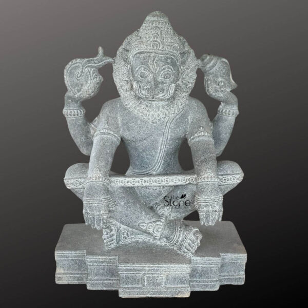 An eighteen-inch-tall intricately carved black granite Sri Narasimha Swamy stone vigraham or murti is depicted in a seated posture directly facing the viewer. The statue is meticulously sculpted from a single block of dark polished stone showcasing the rich crystalline texture and conveying a powerful divine presence. Lord Narasimha the fierce man-lion avatar of Vishnu is seated in a yogic pose upon a tiered pedestal with his left leg folded and his right foot resting on the base. His formidable lion-like face framed by a full stylized mane bears an expression of stern benevolence with wide eyes a powerful snout and a defined mouth. The Narasimha Swamy vigraham has four arms adorned with carved bracelets armlets and shoulder ornaments. In his upper right hand he holds a chakra and in his upper left hand he holds a shankha both symbolic attributes of Vishnu. His lower right hand is raised in the abhaya mudra a gesture of fearlessness and protection while his lower left hand rests gently on his left knee. The statue is detailed with a sacred thread across his chest multiple strands of necklaces and a decorative belt around his waist. The carving captures the fine texture of his mane the folds of his garment and the ornate details of his jewelry and pedestal. This exquisite eighteen-inch black granite Narasimha statue is a perfect representation of traditional South Indian temple art expertly crafted to serve as a revered murti for worship or a significant spiritual artifact within a home shrine. The high-quality black granite material ensures the sculpture's durability and timeless beauty making it a lasting addition to any sacred space. The profound detail in the carving from the fierce yet compassionate expression to the symbolic attributes and ornaments showcases the skill of the artisans and makes this vigraham an ideal choice for devotees seeking a powerful form of the Lord for darshan and spiritual connection. The compact eighteen-inch size allows for placement on a home altar or pedestal bringing the divine energy of Sri Narasimha into the daily lives of the faithful.