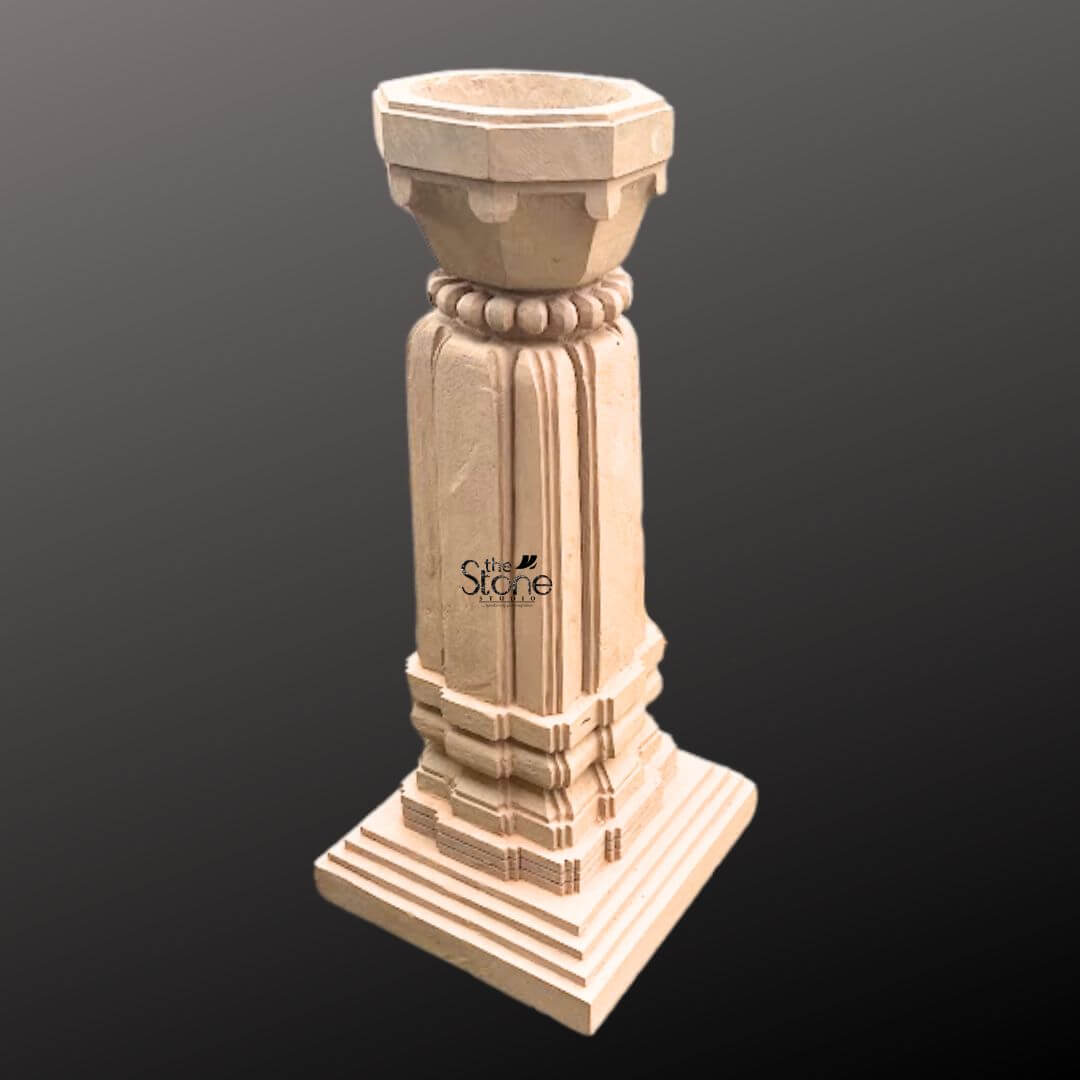 A three-quarter view photograph of a magnificent Stone Tulsi Stand with Planter hand-carved from a single block of warm brown sandstone. Standing approximately 3 feet tall this traditional Indian structure is meticulously crafted to house the sacred Tulsi plant. The entire unit rests upon a substantial multi-tiered square base providing excellent stability and a sense of grounded grandeur perfect for a garden courtyard or sacred space within a home. The main body of the stand features a beautifully proportioned square column adorned with deep vertical fluting on all four faces creating elegant lines that enhance its architectural presence. The sandstone surface displays subtle variations in color and grain highlighting the authentic artisanal quality of the hand-carving process. The precision of the work reflects a deep respect for traditional Indian stonework and temple architecture. Crowning the column is a beautifully shaped flared planter bowl designed to hold the sacred soil and plant. This upper section transitions smoothly from the square column to a circular form featuring a decorative band of carved bead-and-reel molding just below the rim. The overall form is a harmonious blend of functionality and aesthetic grace making this sandstone Tulsi stand a revered and beautiful focal point for any article discussing the significance of the Tulsi plant.