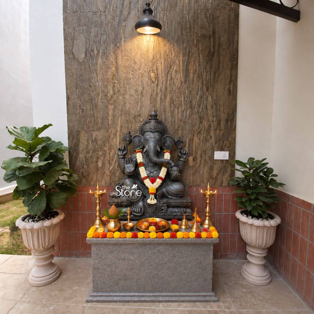 A beautifully composed photograph captures a Traditional Ganesha Stone Statue enshrined in a dedicated home puja space. The central focus is the 2.5ft Blackstone Ganesha meticulously hand-carved from a single piece of deep black stone. The statue depicts Lord Ganesha in a seated posture adorned with a vibrant fresh flower garland of red and white blossoms that contrasts with the dark stone. His form is detailed with traditional jewelry and carvings and he is seated upon a simple rough-hewn granite pedestal that serves as a home altar. The altar area is arranged with meticulous care for traditional worship. The pedestal is covered by a thick border of bright yellow marigold flowers and interspersed red and yellow blossoms. Placed on the altar are several essential ritual items including two tall lit brass oil lamps on either side a central brass pot with a coconut and mango leaves and a collection of small metal bowls containing offerings and powders. This setup demonstrates the active spiritual use of the Ganesha stone statue in a domestic setting. The backdrop for this shrine enhances the traditional aesthetic. The statue is set against a feature wall of textured rustic brown stone cladding which is illuminated from above by a single modern black pendant light casting a warm focused glow on the deity. The wall transitions to plain white plaster on the left and a section of reddish-brown brick tile on the right. Flanking the entire altar setup are two large ornate stone planters each holding a healthy Fiddle Leaf Fig plant adding life and symmetry to the sacred space. The floor is paved with light-colored stone tiles creating a clean and grounded foreground.