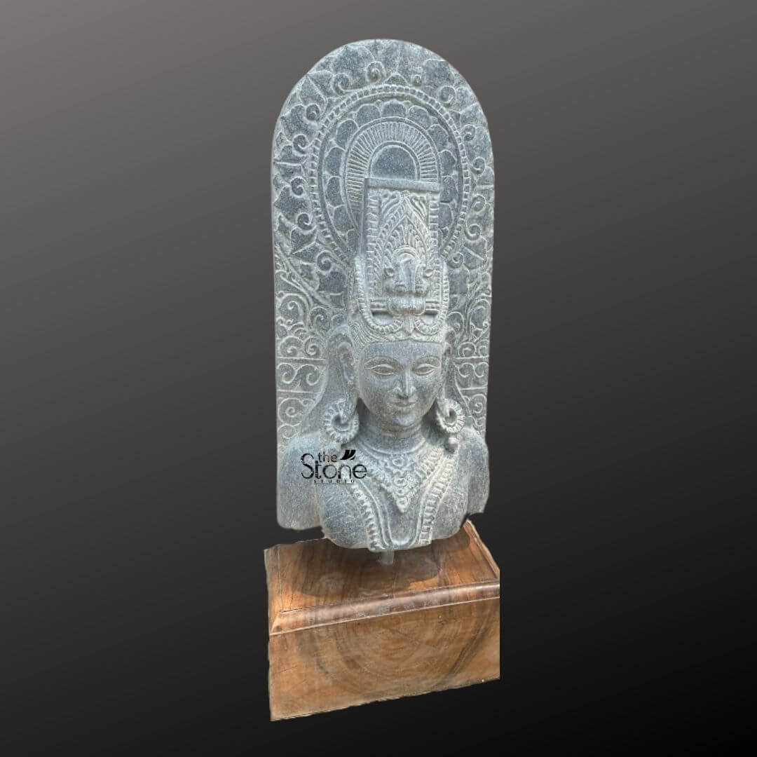 A magnificent thirty-inch hand-carved Traditional Lord Shiva Stone Bust stands as the revered centerpiece against a dark neutral gradient background. The sculpture meticulously chiseled from a single block of deep black stone exhibits a rich aged texture with natural variations and a weathered patina that underscores its antique character. Lord Shiva is portrayed with a serene contemplative facial expression characterized by eyes gently closed in meditation arched eyebrows and a subtle composed smile that conveys a profound sense of peace and divine wisdom. The bust features elaborate ornamentation including large circular earrings and multiple layered necklaces with intricate pendants that rest upon his chest all hand-carved with precision. His hair is intricately styled in a traditional jata forming a high crown adorned with delicate carvings of a crescent moon a serpent and a central floral motif with further details of coiled locks falling onto his shoulders. Behind the deity's head a richly carved ornate prabhavali rises featuring detailed symmetrical floral and scrollwork patterns that frame his divine form. The craftsmanship showcases the traditional South Indian temple art style with deep undercuts and high relief that create a profound sense of depth and reverence. The entire sculpture rests securely on a dark polished wooden plinth. This exquisite thirty-inch hand-carved Traditional Lord Shiva black stone bust is a perfect testament to the timeless devotion and artistic skill of traditional Indian artisans. The large scale of the bust combined with the deep black stone and the profound meditative expression makes it an awe-inspiring focal point for any spiritual space or home mandir. The detailed iconography from the elaborate headdress to the intricate jewelry and the ornate prabhavali ensures this antique-style murti meets the highest aesthetic and spiritual requirements for those seeking a significant and beautiful representation of Lord Shiva. The natural color of the stone and the profound depth of the carving convey a sense of sanctity and spiritual presence making it a lasting addition to any sacred environment.