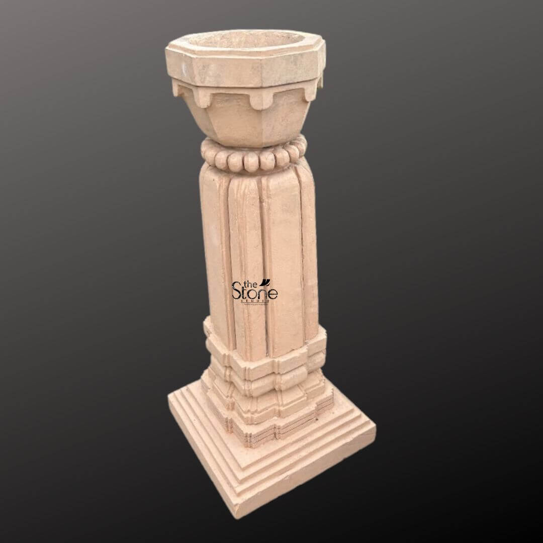 A three-quarter view photograph of a magnificent Traditional Tulsi Vrindavan in Stone hand-carved from a single block of warm brown sandstone. Standing approximately 3 feet tall this sacred planter is meticulously crafted in a classic architectural style designed specifically for housing the revered Tulsi plant. The entire structure rests upon a substantial multi-tiered square base providing stability and a sense of grounded reverence suitable for a home courtyard or garden. The main body of the Vrindavan features a square column adorned with deep elegant vertical fluting on all four faces creating clean lines that enhance its architectural presence. The natural texture and warm hues of the sandstone are beautifully showcased reflecting the authentic artisanal quality of the hand-carving process. The precision and symmetry of the work highlight the skill involved in creating this traditional Indian stone art. Crowning the column is a beautifully proportioned flared planter bowl designed to hold the sacred soil and plant. This upper section transitions smoothly from the square column to a circular form featuring a decorative band of carved bead-and-reel molding just below the rim. The overall design is a harmonious blend of structural integrity and delicate artistry making this sandstone Vrindavan a revered and beautiful focal point for any article discussing the tradition of Tulsi worship.