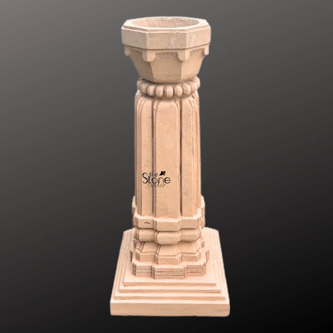 A detailed three-quarter view photograph of a magnificent Tulsi Pot in Stone hand-carved from a single block of warm brown sandstone. This traditional Indian planter standing approximately 3 feet tall is meticulously designed to house the sacred Tulsi plant. The entire structure rests upon a substantial multi-tiered square base providing excellent stability and a sense of grounded grandeur perfect for a home courtyard or garden setting. The main body of the pot features a beautifully proportioned square column adorned with classic vertical fluting on all four faces creating elegant lines that enhance its architectural presence. The sandstone surface displays subtle variations in color and grain highlighting the authentic artisanal quality of the hand-carving process. The precision of the work reflects a deep respect for traditional Indian stone carving techniques. Crowning the column is a beautifully shaped flared planter bowl designed to hold the sacred soil and plant. This upper section transitions smoothly from the square column to a circular form featuring a decorative band of carved bead-and-reel molding just below the rim. The overall form is a harmonious blend of functionality and aesthetic grace making this sandstone Tulsi pot a revered and beautiful focal point for any article discussing the cultural and spiritual significance of the plant.