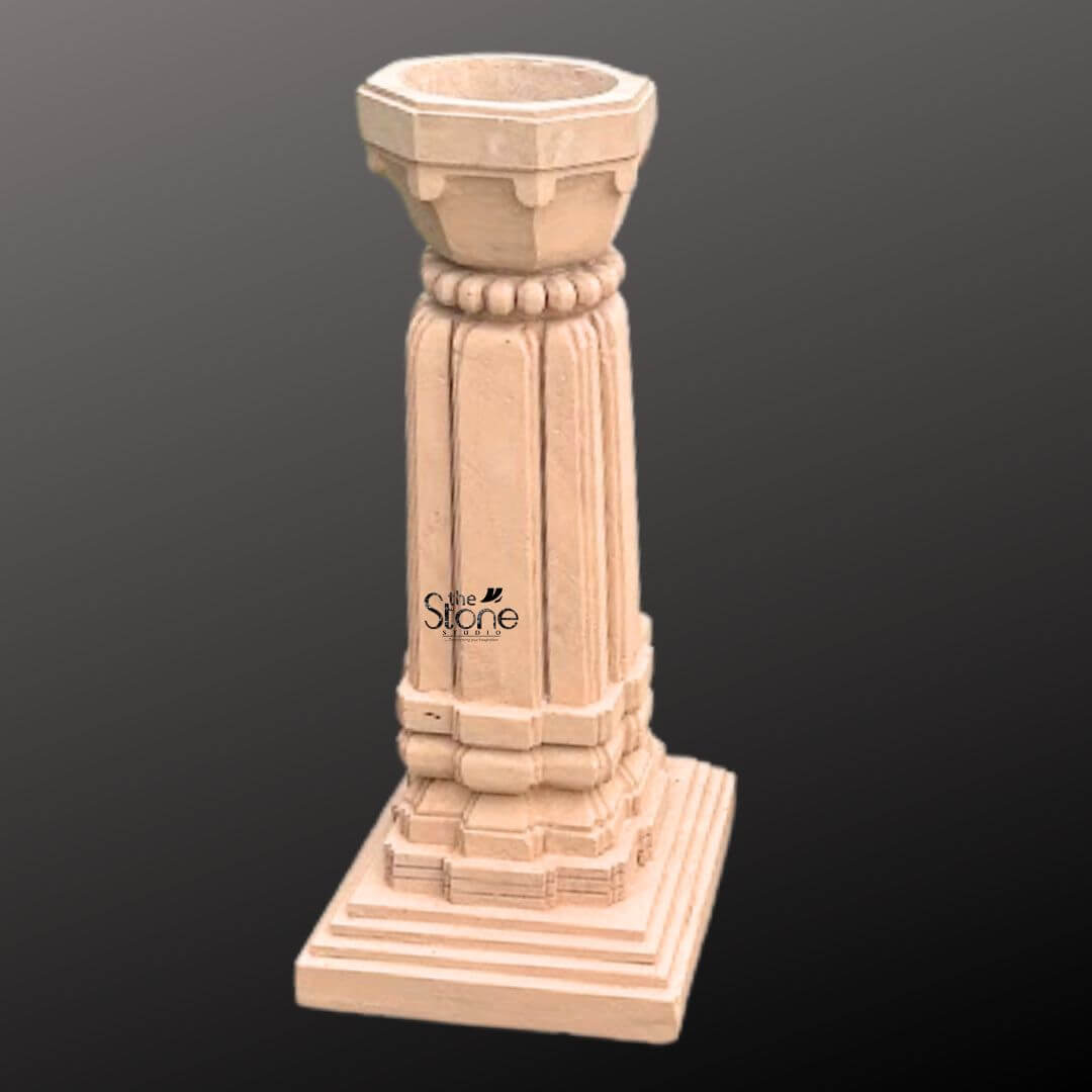 A three-quarter view photograph of a magnificent Tulsi Vrindavan Stone Planter hand-carved from a single block of warm brown sandstone. Standing approximately 3 feet tall this traditional Indian planter is meticulously crafted in a classic architectural style designed specifically for housing the sacred Tulsi plant. The entire structure rests upon a substantial stepped square base providing excellent stability and a sense of grounded grandeur perfect for a garden or courtyard setting. The main body of the planter features a beautifully proportioned square column adorned with classic vertical fluting on all four faces creating elegant lines that enhance its architectural presence. The sandstone surface displays subtle variations in color and grain highlighting the authentic artisanal quality of the hand-carving process. The precision of the work reflects a deep respect for traditional Indian stone carving techniques. Crowning the column is a beautifully shaped flared planter bowl designed to hold the sacred soil and plant. This upper section transitions smoothly from the square column to a circular form featuring a decorative band of carved bead-and-reel molding just below the rim. The overall form is a harmonious blend of structural strength and delicate artistry making this sandstone Tulsi Vrindavan a revered and beautiful focal point for any home or garden.