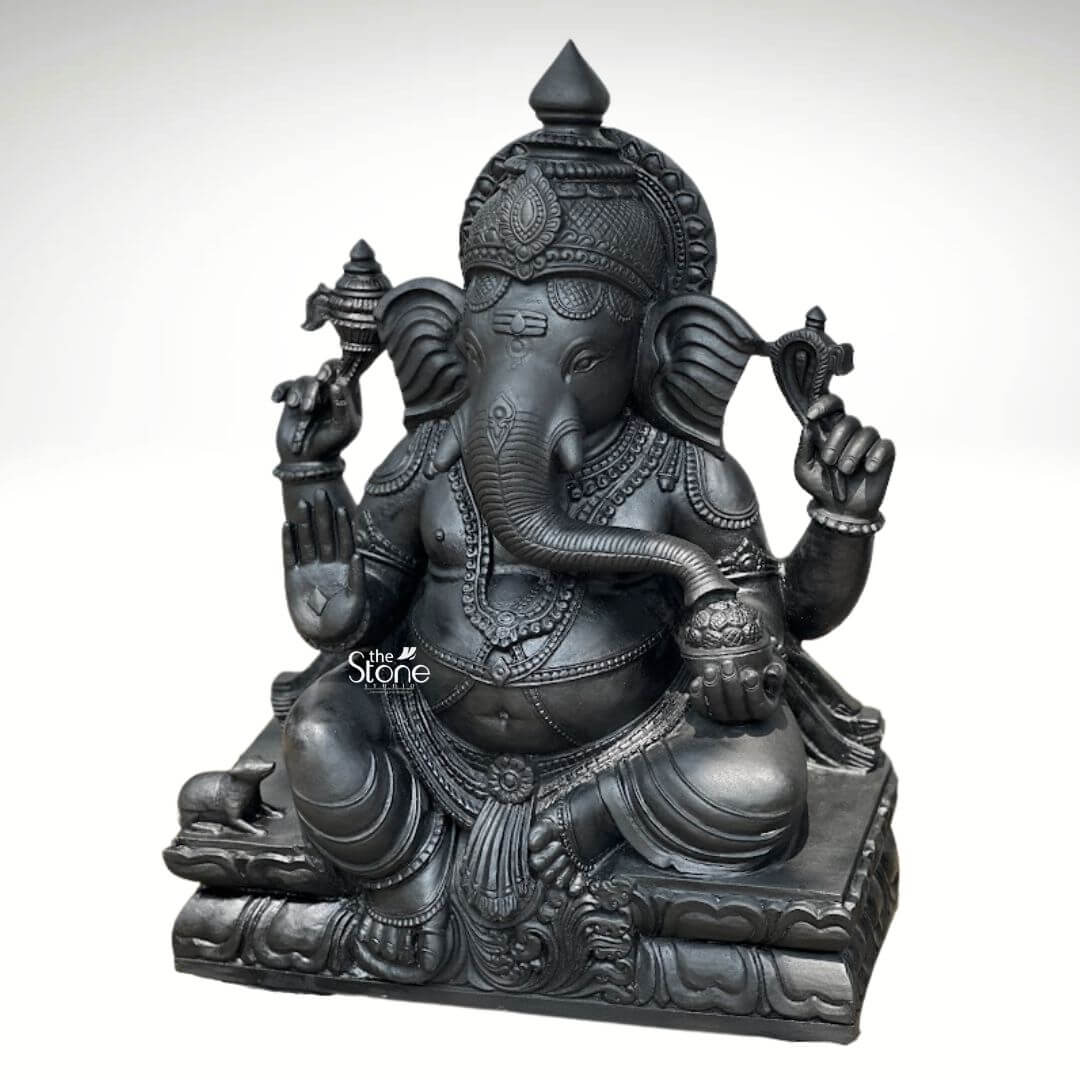 A full three-quarter view photograph of a magnificent Vinayagar Black Stone Statue expertly hand-carved from a single piece of deep black stone. The statue which stands approximately 2.5 feet tall depicts Lord Ganesha in a seated majestic posture. His form is robust and well-proportioned characterized by a smooth dark finish that accentuates the natural texture and intricate details of the stone. The deity is seated on a beautifully carved pedestal designed to resemble stacked lotus petals providing a stable and traditional base. Ganesha is adorned with rich deeply cut ornamentation across his body including multiple necklaces elaborate armbands wristlets and anklets all rendered in the same black stone. He wears a decorated crown and a sacred thread across his chest. He has four arms his upper right hand holds a parashu and his upper left holds a pasha. His lower right hand is raised in the abhaya mudra while his lower left hand cradles a modaka which his gracefully curved trunk is poised to taste. This Vinayagar Black Stone Statue showcases the exemplary skill of traditional Indian stonework. The fine details in the carving, such as the traditional tilak markings on the forehead the large expressive ears and the folds of his garment highlight the spiritual significance and artistic craftsmanship associated with these sacred sculptures. The dark solemn presence of the basalt stone makes this 2.5ft statue a commanding and revered focal point for any article discussing the beauty and tradition of Vinayagar Black Stone Statues.