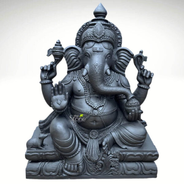 A magnificent Vinayaka Ganapathi Black Stone Statue stands as a profound example of traditional South Indian sculptural art. Hand-carved from a single block of deep blackstone the statue depicts the Hindu deity Ganesha in a traditional seated posture known as the Lalitasana pose with his right leg folded and left leg slightly raised. The dark polished stone surface exhibits a subtle cool sheen under the studio lighting highlighting the smooth texture of the material. The artisan has lavished attention on the intricate details from the ornate crown and multiple necklaces adorning his form to the delicate folds of his flowing dhoti and the individual petals of the lotus throne he occupies. Ganesha is depicted with four arms each holding a symbolic item with great precision his upper right hand holds an ankusha his upper left holds a pasha and his lower right hand is in the Abhaya Mudra bestowing blessings upon devotees. His lower left hand holds a modak resting in his lap with his trunk gently curled to sample the offering. The statue features a prominent finely carved prabhavali behind the deity, adorned with traditional patterns and culminating in a kirtimukha at the apex. His loyal vahana the mouse is seated attentively at his feet to the left completing the divine composition. This black stone Vinayaka Ganapathi statue is an ideal size for a prominent home shrine a temple sanctum or as a significant decorative piece. The substantial weight and durable stone material ensure its longevity making it a lasting investment for spiritual practice or cultural decor. The photograph against a plain white background emphasizes the statue's three-dimensional quality and the artisan's meticulous work on the serene facial expression and decorative elements perfectly illustrating the grace and cultural richness that a well-crafted black stone Ganesha statue brings to any space.