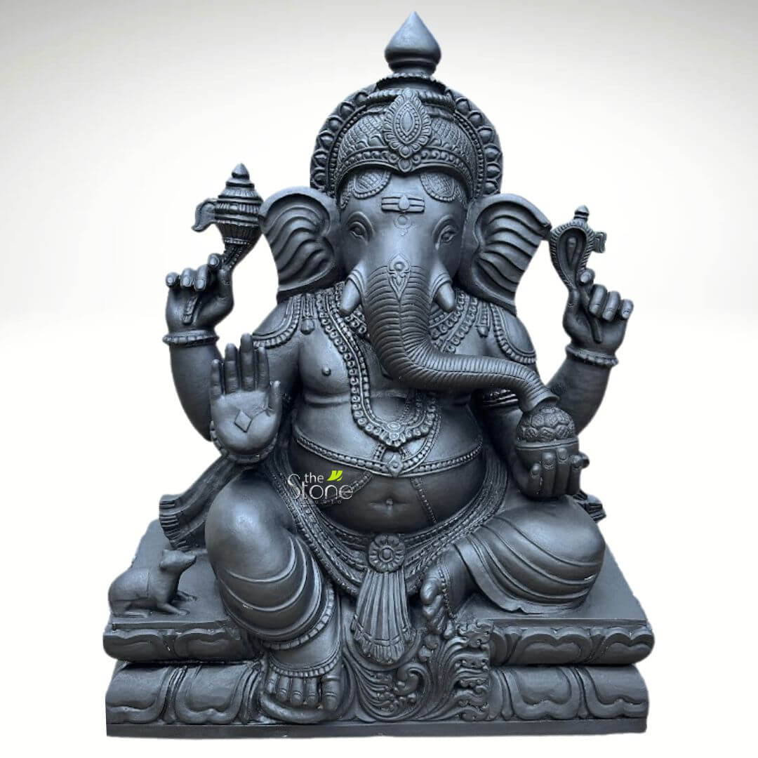 A magnificent Vinayaka Ganapathi Black Stone Statue stands as a profound example of traditional South Indian sculptural art. Hand-carved from a single block of deep blackstone the statue depicts the Hindu deity Ganesha in a traditional seated posture known as the Lalitasana pose with his right leg folded and left leg slightly raised. The dark polished stone surface exhibits a subtle cool sheen under the studio lighting highlighting the smooth texture of the material. The artisan has lavished attention on the intricate details from the ornate crown and multiple necklaces adorning his form to the delicate folds of his flowing dhoti and the individual petals of the lotus throne he occupies. Ganesha is depicted with four arms each holding a symbolic item with great precision his upper right hand holds an ankusha his upper left holds a pasha and his lower right hand is in the Abhaya Mudra bestowing blessings upon devotees. His lower left hand holds a modak resting in his lap with his trunk gently curled to sample the offering. The statue features a prominent finely carved prabhavali behind the deity, adorned with traditional patterns and culminating in a kirtimukha at the apex. His loyal vahana the mouse is seated attentively at his feet to the left completing the divine composition. This black stone Vinayaka Ganapathi statue is an ideal size for a prominent home shrine a temple sanctum or as a significant decorative piece. The substantial weight and durable stone material ensure its longevity making it a lasting investment for spiritual practice or cultural decor. The photograph against a plain white background emphasizes the statue's three-dimensional quality and the artisan's meticulous work on the serene facial expression and decorative elements perfectly illustrating the grace and cultural richness that a well-crafted black stone Ganesha statue brings to any space.