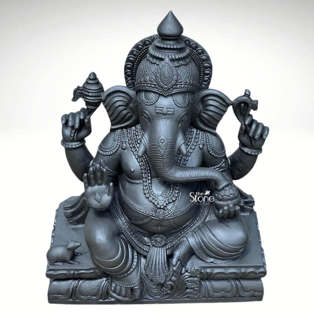 A full three-quarter view photograph of a magnificent Blackstone Ganesha statue meticulously carved and standing 2.5ft tall. The deity revered as Vinayaka Ganapathi Murti is depicted in a traditional seated posture known as lalitasana with one leg folded and the other resting comfortably. The statue is crafted from a single piece of deep black stone possessing a smooth matte finish that accentuates the intricate details and the natural dark texture of the basalt. Lord Ganesha is adorned with elaborate deeply carved ornaments including multiple necklaces armlets wristlets and anklets all rendered in the same black stone. He wears a richly decorated crown and a sacred thread across his chest. He is four-armed his upper right hand holds a parashu and his upper left holds a pasha. His lower right hand is raised in the abhaya mudra while his lower left hand cradles a modaka which his gracefully curved trunk is poised to taste. The elephant head features traditional tilak markings on the forehead. The statue is seated upon a substantial tiered pedestal carved to resemble stacked lotus petals providing a stable and elegant foundation. At the lower left of the pedestal near Ganesha's foot a small intricately carved mooshik his vahana is depicted in a reverent pose looking up towards the deity. The overall composition highlights the divine symmetry and rich iconography of this masterwork making it a quintessential example of a Vinayaka Ganapathi Murti suitable for spiritual spaces and art collections. The dark solemn presence of the black stone imparts a sense of ancient tradition and reverence.