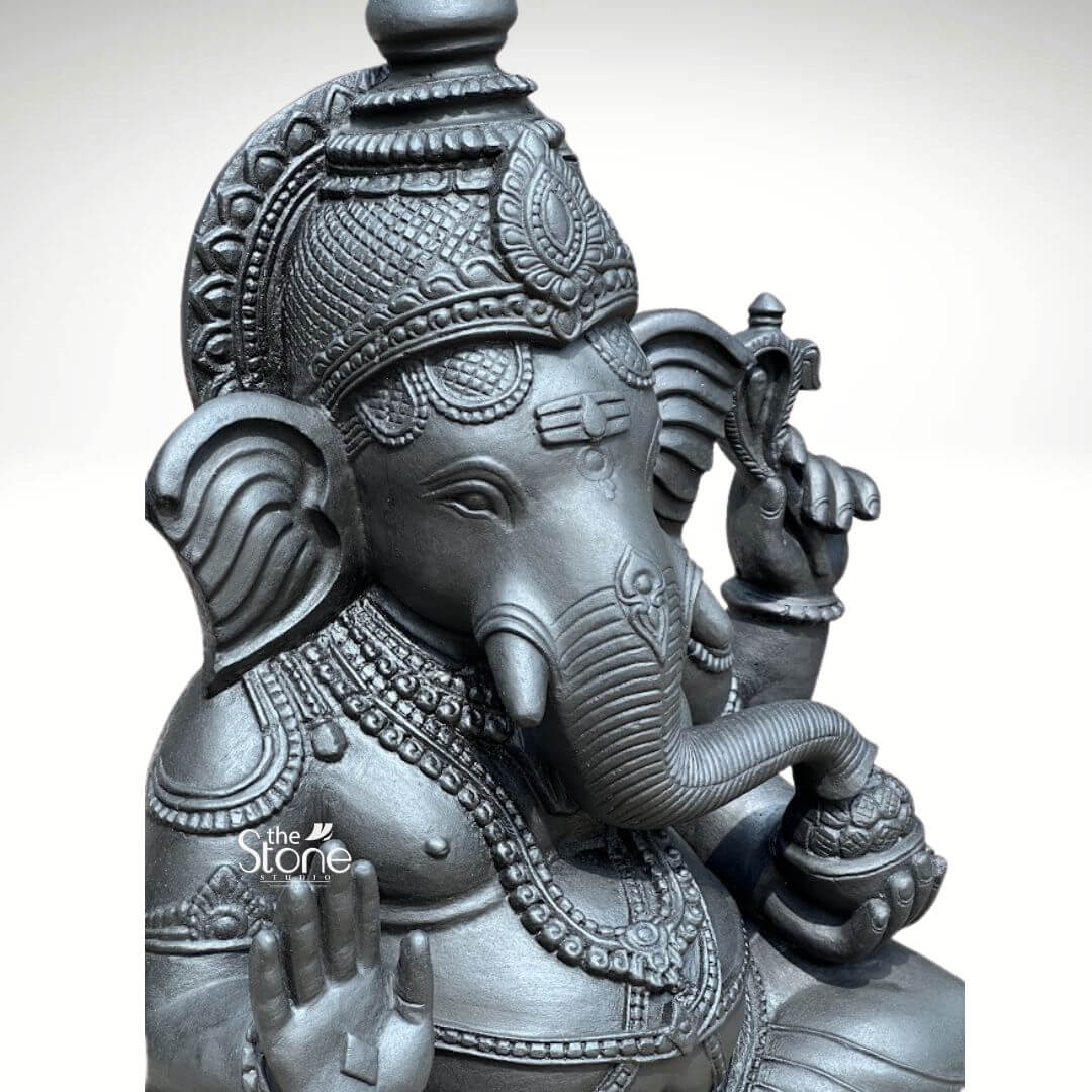A close-up three-quarter view photograph focusing on the upper portion of a magnificent Blackstone Ganesha statue identified as a 2.5ft Vinayaka Stone Vigraham. The deity is depicted in a seated posture meticulously carved from a single block of deep dark black stone which gives the sculpture a solemn and ancient appearance. The smooth matte finish of the basalt stone highlights the natural texture and the exceptional precision of the hand-carving showcasing the divine form. The elephant-headed god is adorned with elaborate deeply-cut ornamentation that covers his chest arms and head. He wears a richly decorated multi-tiered crown and multiple strands of intricate necklaces and armlets. A sacred thread is visible across his torso all rendered in the same dark stone with detailed patterns. His face exudes serenity with traditional tilak markings on his forehead and large expressive ears. Lord Ganesha is shown with four arms his upper right hand holds a parashu and his upper left hand holds a pasha. His lower right hand is visible in the foreground raised in the abhaya mudra while his trunk curves gracefully to the left to hold and taste a modaka held in his lower left hand. The photograph captures the intricate details of the jewelry the texture of the skin and the spiritual grace of the sculpture making it an exemplary Vinayaka Stone Vigraham for worship or art appreciation.